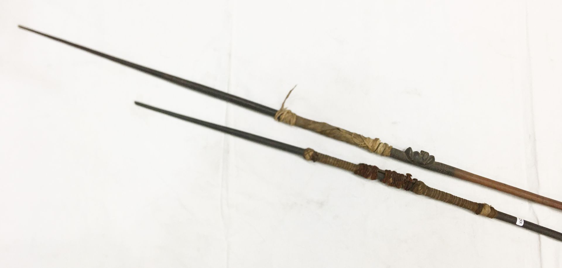 Null 两支Kanake硬币长矛的拍品，一支有雕刻头像的装饰，另一支是残缺不全。木材、纤维和狗鱼毛。长：237厘米