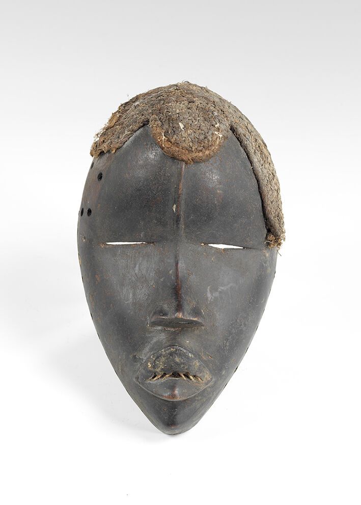 Null 一个带有黑色铜锈和纤维的硬木面具，表现了一张椭圆形的人脸，充满了宁静的气息。额头有中间的肋骨，眼睛有细缝，嘴巴很好。DAN，象牙海岸。高度：21厘米。