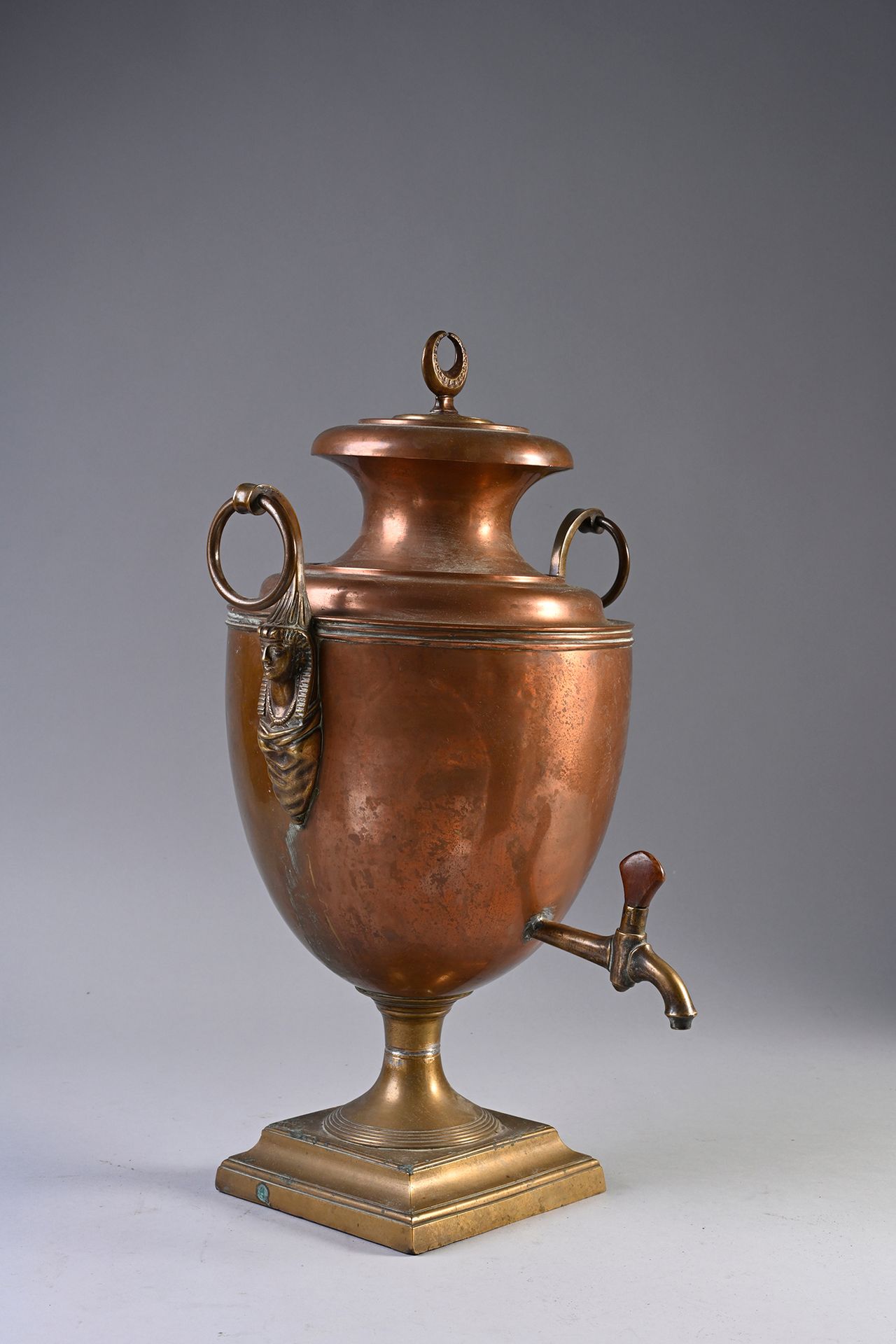 Null 优雅的红铜茶炊。
俄罗斯作品，19世纪初。
瓮的形状，安放在一个黄铜的方形底座上，侧面有镂空的环形抓手，最后是埃及风格的妇女头像，上部有一个月牙。包括&hellip;