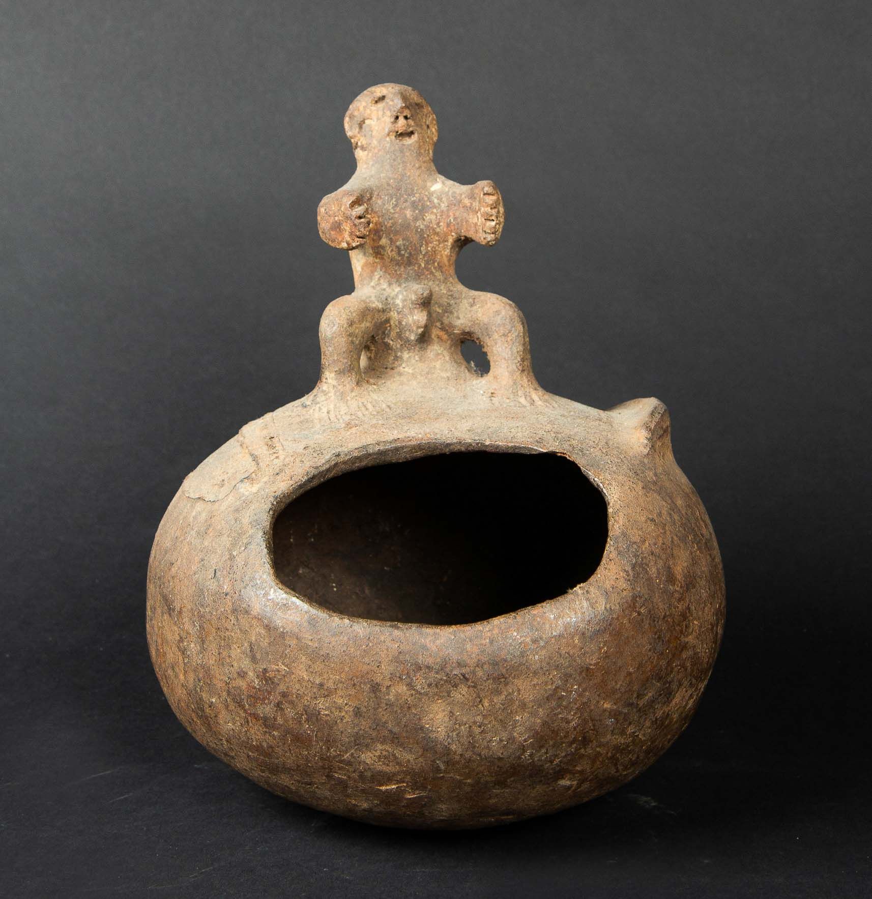 Null 一个米色和棕色赤土的古代祭祀容器，有缺失的部分和左边的突起。它显示了一个坐着的政要，双手比例有力。尼日利亚，伊兹。高：21厘米