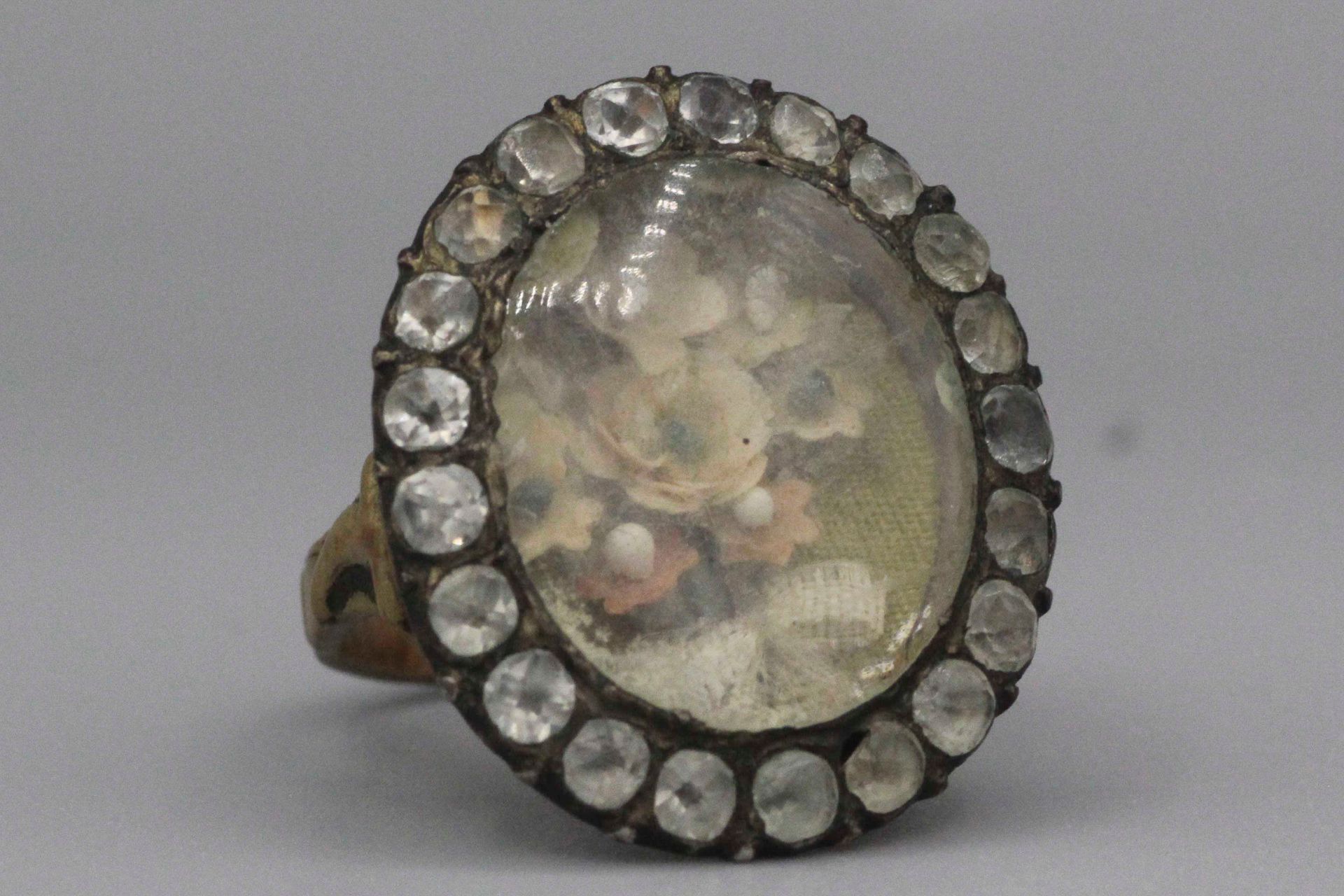 Null Ring aus Metall mit Blumen unter Glas in einer Umrandung aus Rheinsteinen.