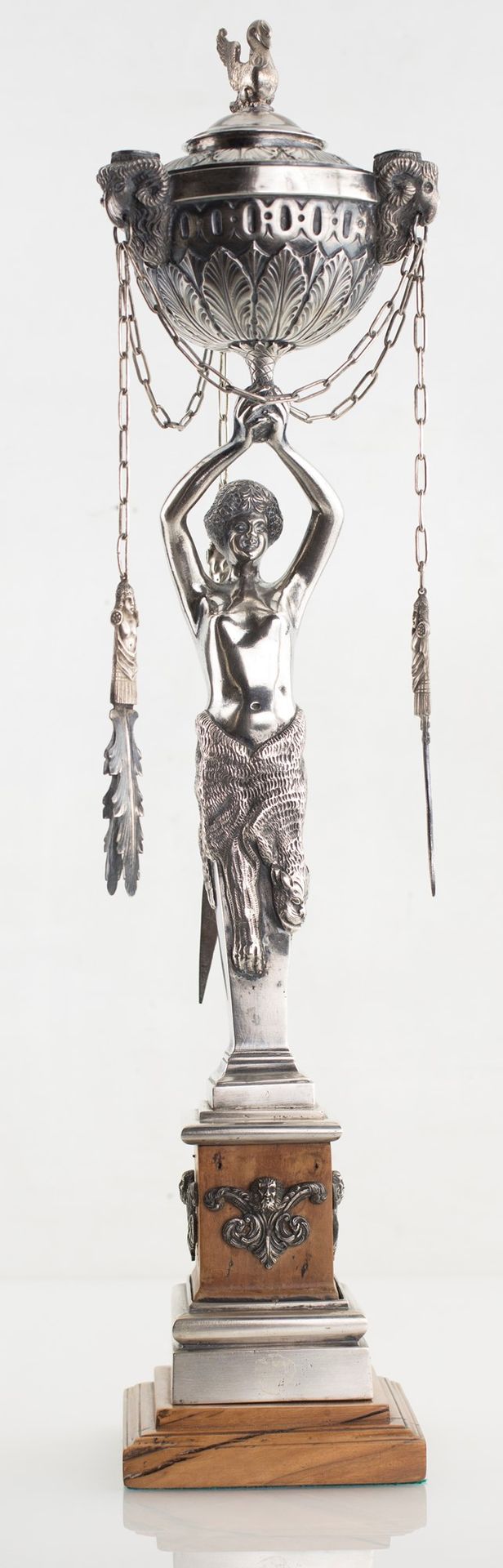 Silver oil lamp, 19th century 由一个豆荚组成，豆荚上有三个带公羊头的水口，由一个部分包裹在狮子皮中的女性荷尔蒙支撑；方形底座