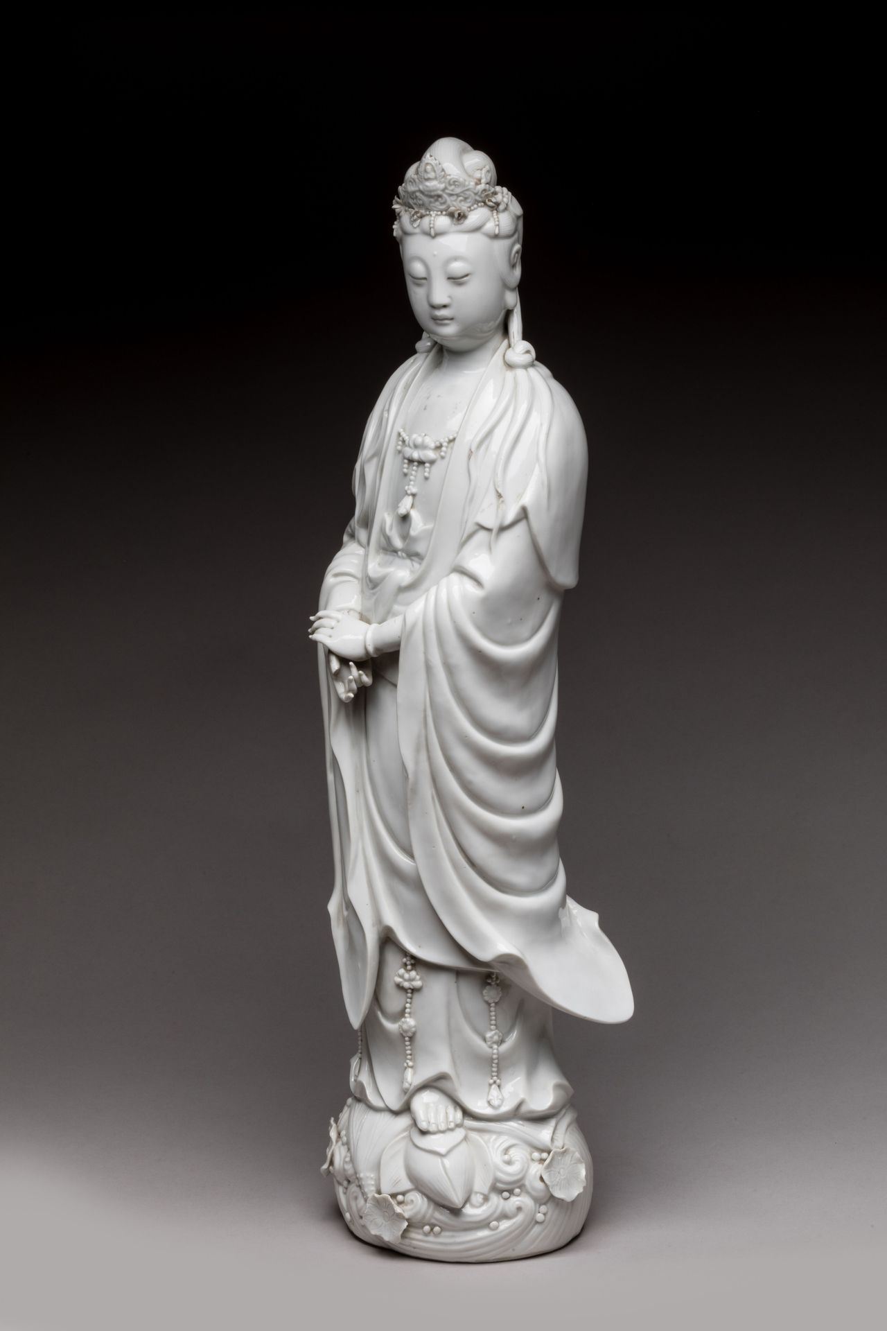 Null 
中国 - 20世纪初 


中国白釉观音瓷像，站在波浪形的莲花上，双手合十，戴着莲花胸饰，头饰有佛像的头饰。 


高度55.5厘米（事故）。 

&hellip;