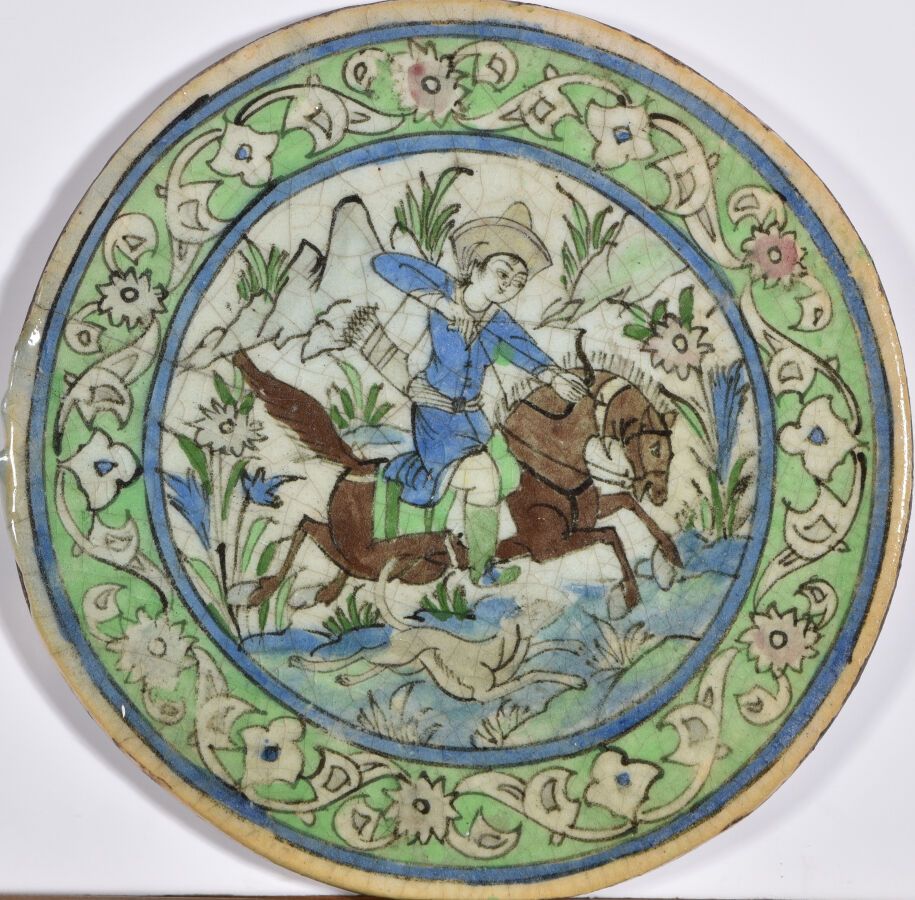 Null IRAN Kadjar 约1920年。

圆形陶瓷瓦片上装饰着一个骑手发射弓箭和一只狗在风景中奔跑，周围是绿色背景上的风格化花朵楣。

D.36厘米。&hellip;