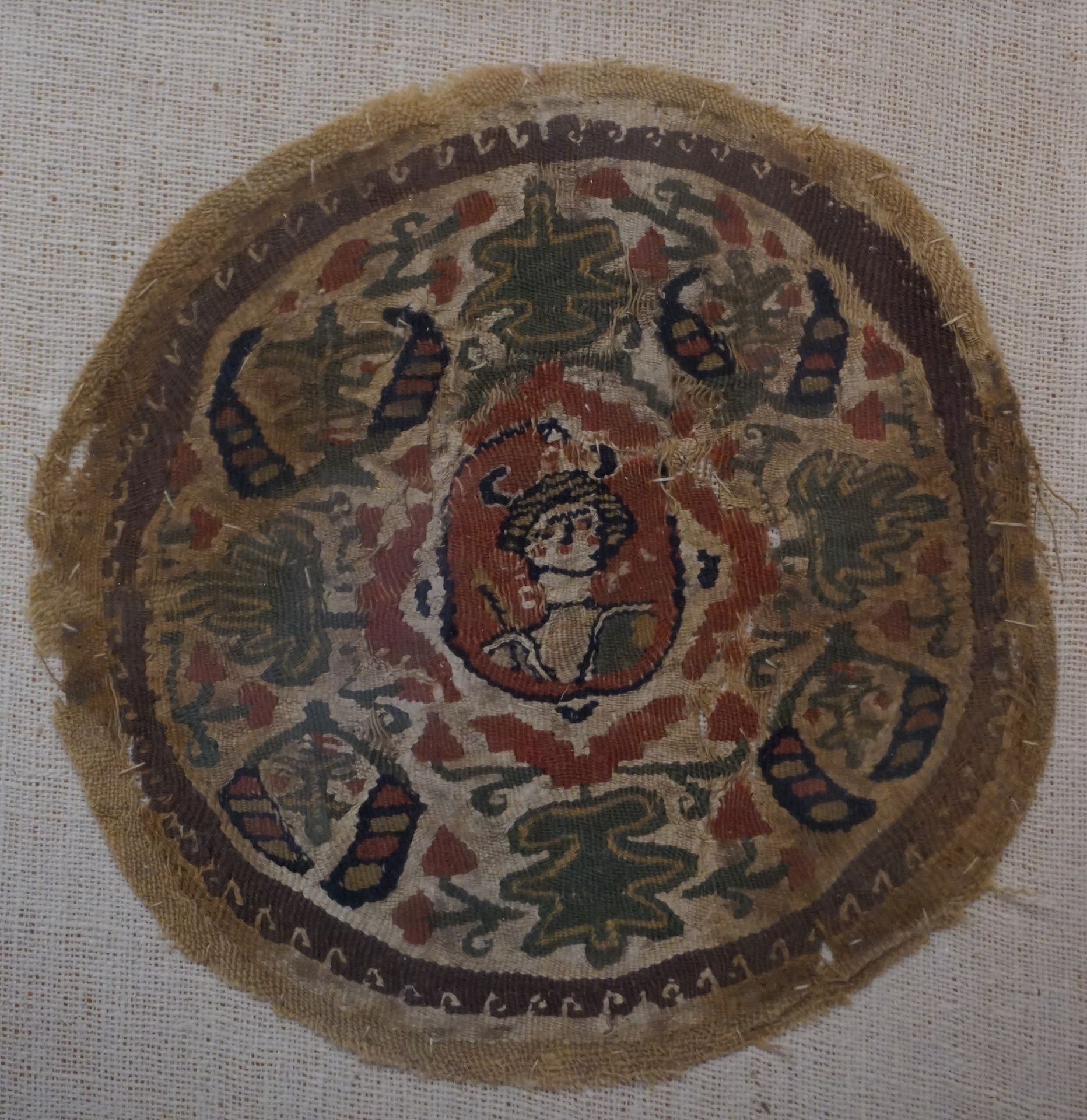 Null 圆形奖章表现了一个半身像的轮廓，并以风格化的植物图案为框架。绿色、红色、黑色和米色的亚麻布。事故和污点。埃及，6-8世纪。直径：12.5厘米