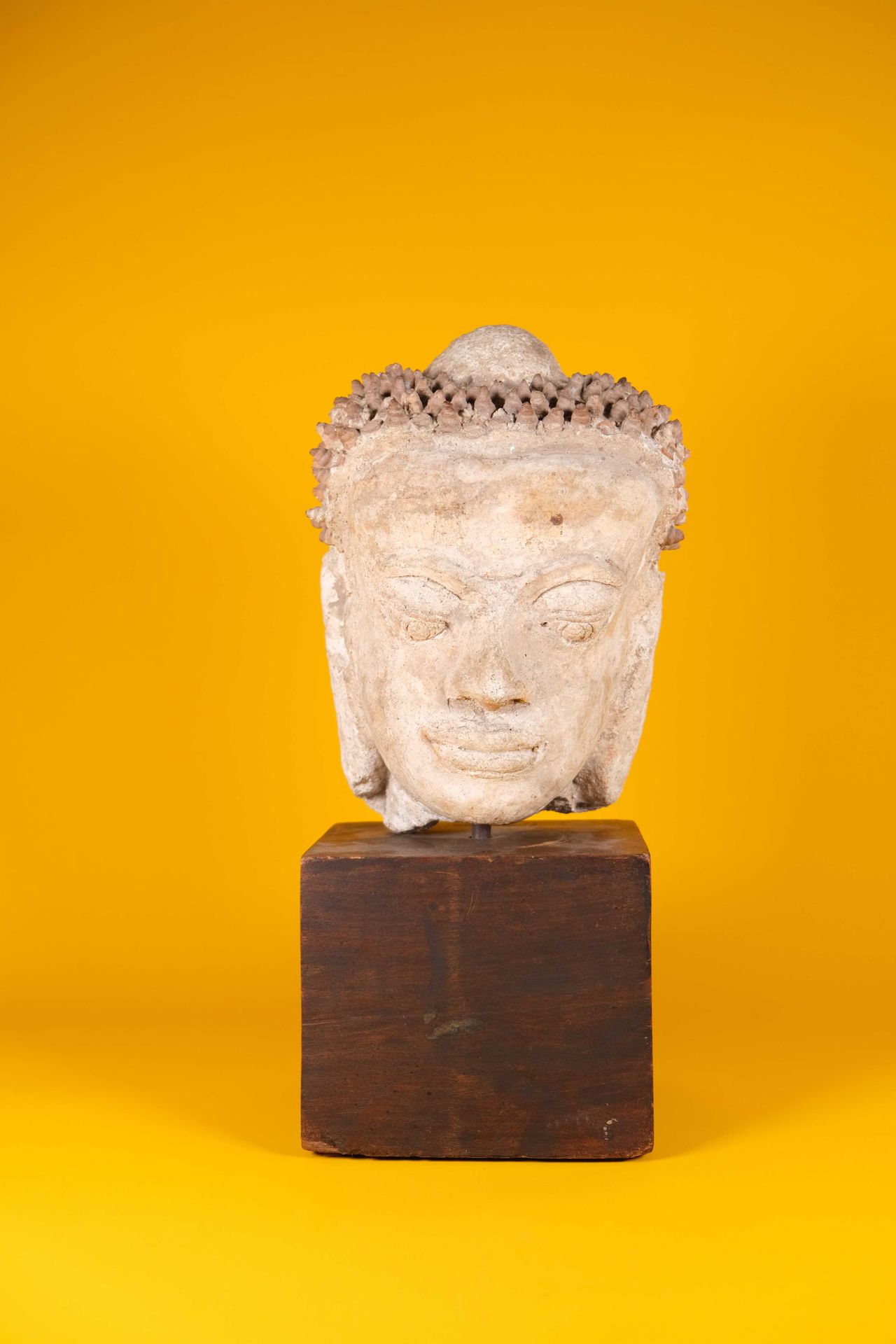 Buddha head in stucco, the face serene, the forehead marked' with the urna,  the hair in curls and covering the ushnisha, wooden base. Thailand,  Haripunchai, 13th century. H: 27 cm (visible lacks)