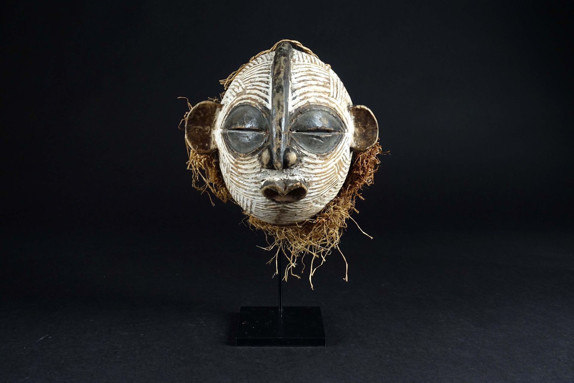 Culture Luba. 小型Kifwebe面具，圆形，有一个孵化的脸，突出的耳朵和一个心形的嘴。雕刻的木头上有青铜色和白色颜料，还有酒椰叶。刚果民主共和国。&hellip;