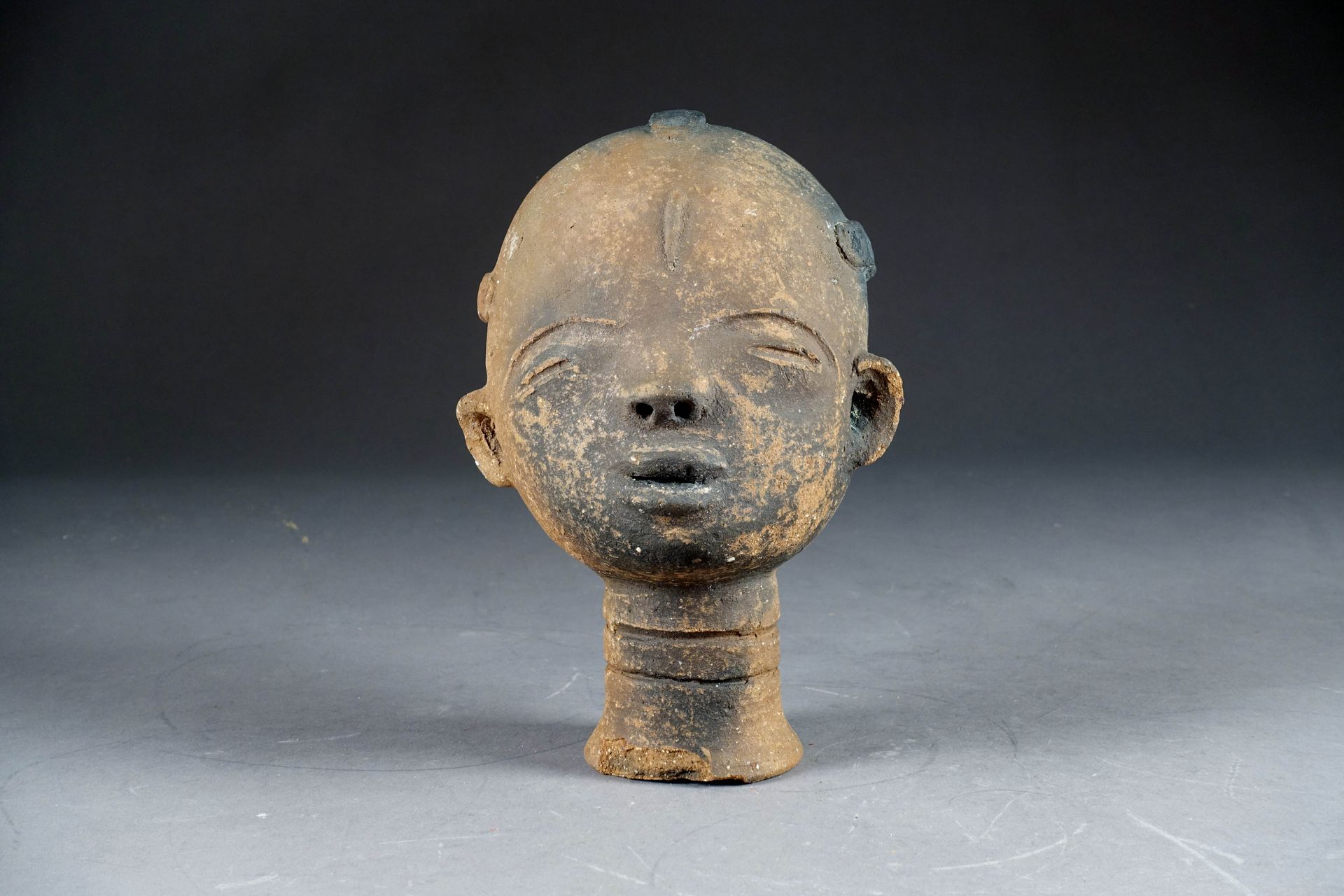 Culture Akan. 风格化的纪念性头像，半闭着眼睛，额头上有疤痕。带有火的痕迹的赤土。加纳。高度：24厘米。
