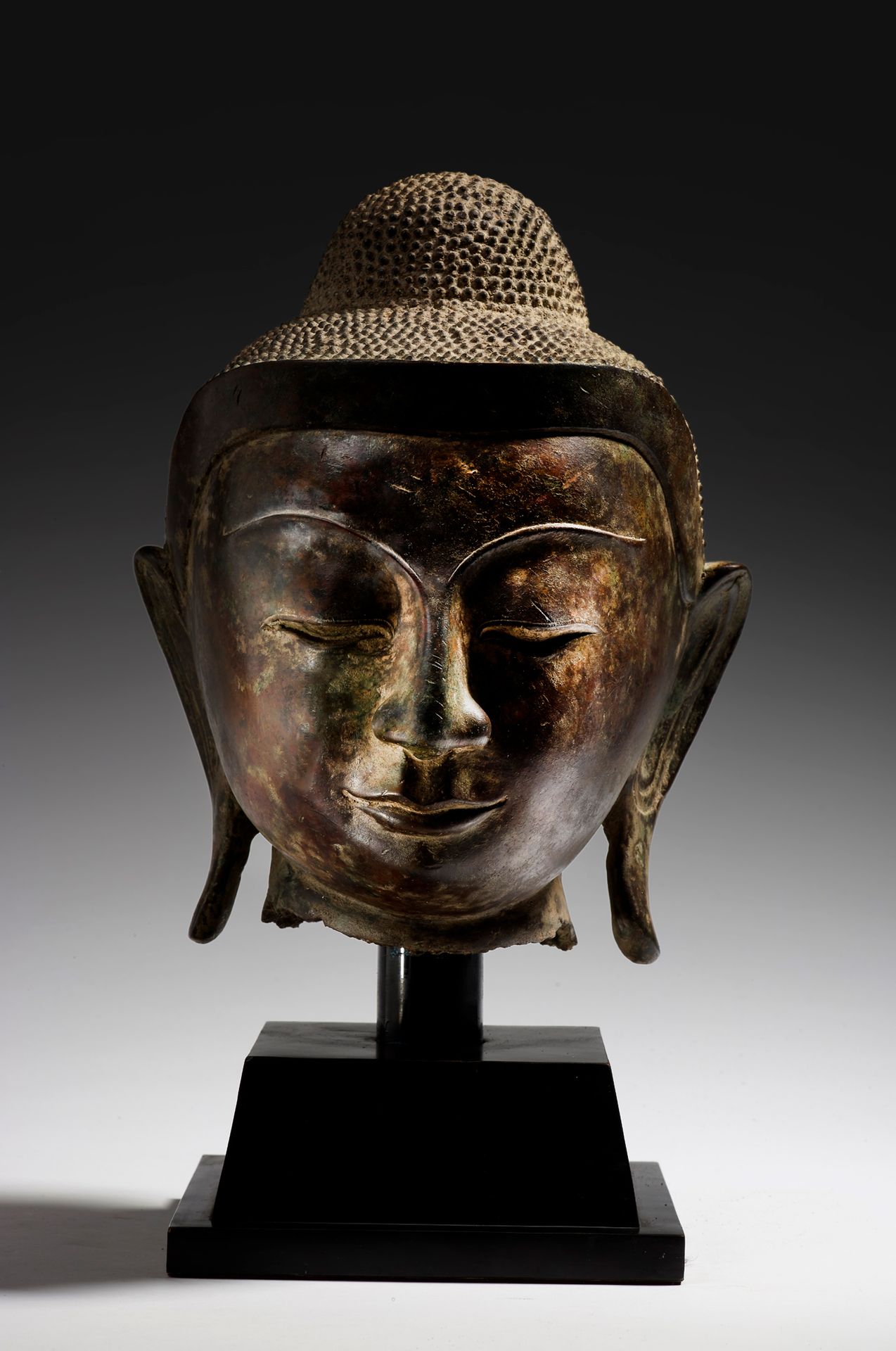 Large Buddha head in bronze with green patina, eyes half closed and  smiling, hair gathered in small tight curls topped by the usnisa. BURMA,  Mandalay - 19th century H. 30 cm (scratches, accidents)