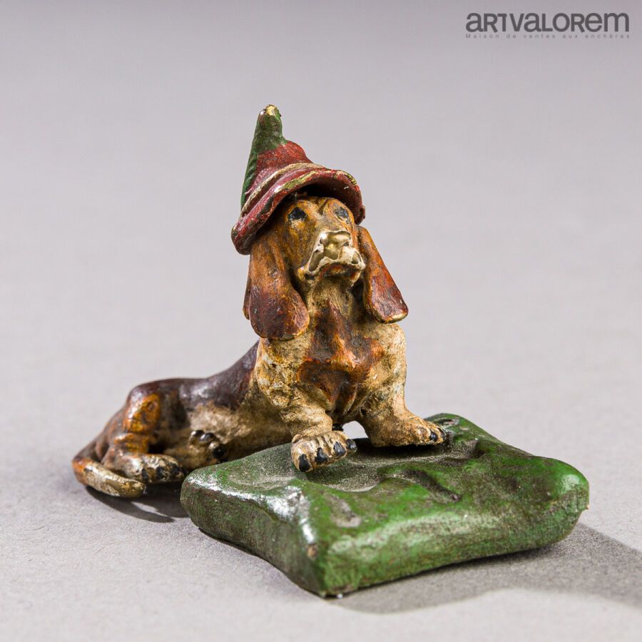 Null 一尊多色铜像，是一只躺着的巴吉特猎犬，戴着红色的羽毛帽，前腿搭在一个绿色的垫子上。
维也纳，20世纪。
高度：3.5厘米 - 长度：5厘米 - 深度：&hellip;