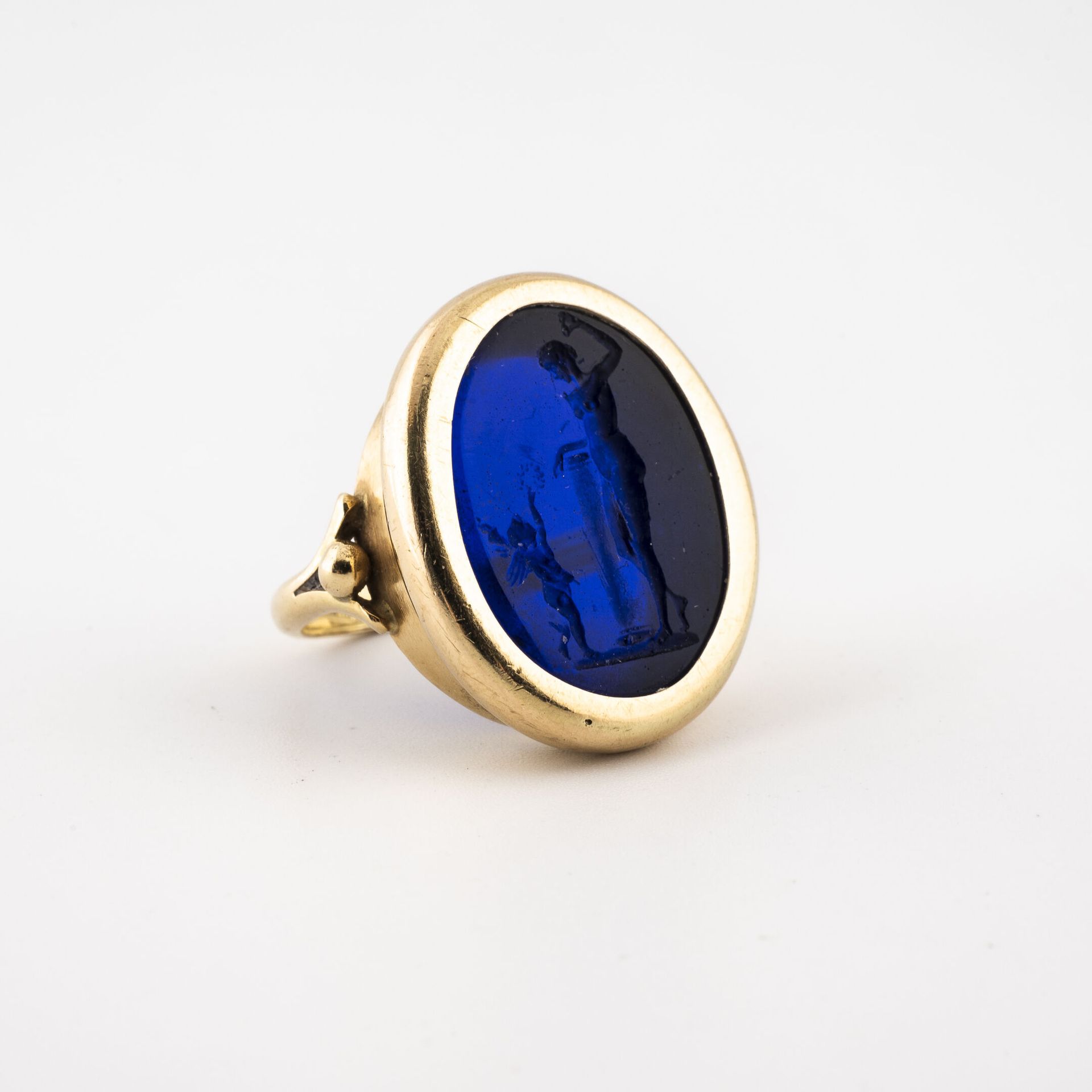 Null 一枚黄金（750）戒指，椭圆形的顶部镶嵌着蓝色的玻璃凹版，描绘了一个靠在基座上的裸体女人和一个拿着皇冠的普特人。

毛重：8.3克。- 手指大小：51&hellip;