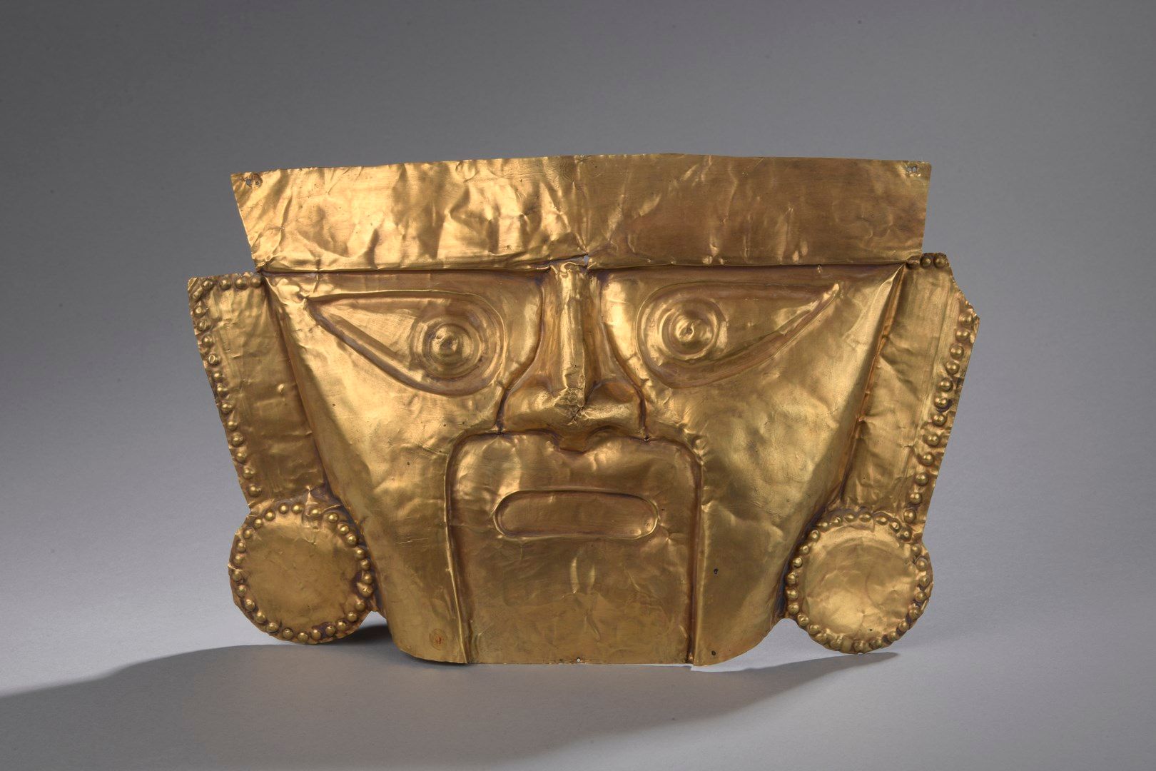 Null 邪教面具显示一个领主的脸，他的耳朵上带着两个圆形的坦巴。

切割和压印的金箔。南美洲。

高度：20.2厘米 - 长度：32厘米

鼻部凹陷，小事故，&hellip;