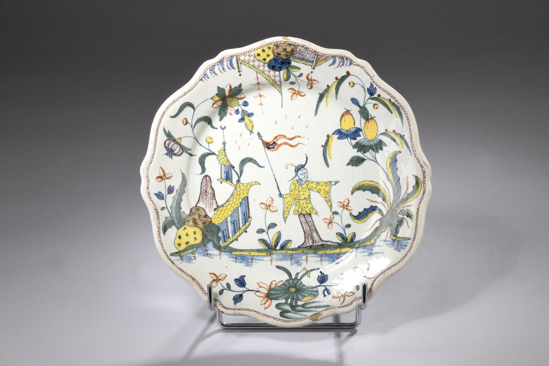 Null 鲁昂 - 18世纪
陶器盘子，边缘有轮廓，有多色装饰，一个中国人在水生植物中间的宝塔附近拿着一面旗子。
边缘有一个小裂缝和缺口。
直径24,8厘米