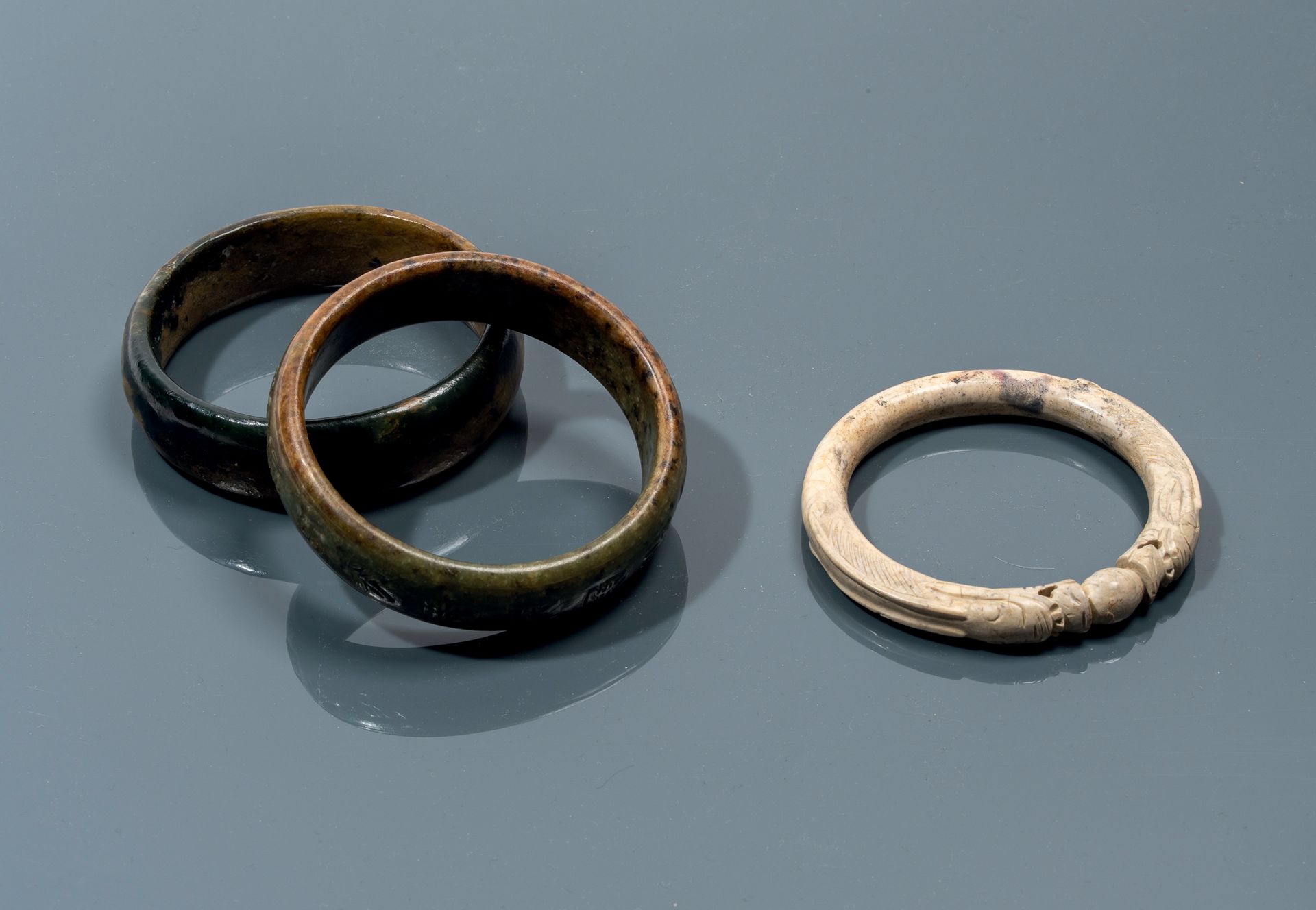 CHINE - Epoque MING (1368 - 1644) 
三个软玉手镯，一个是鸡骨形的双龙合璧的珠子，一个是青瓷棕色的带古文字装饰的手镯，一个是黑棕&hellip;