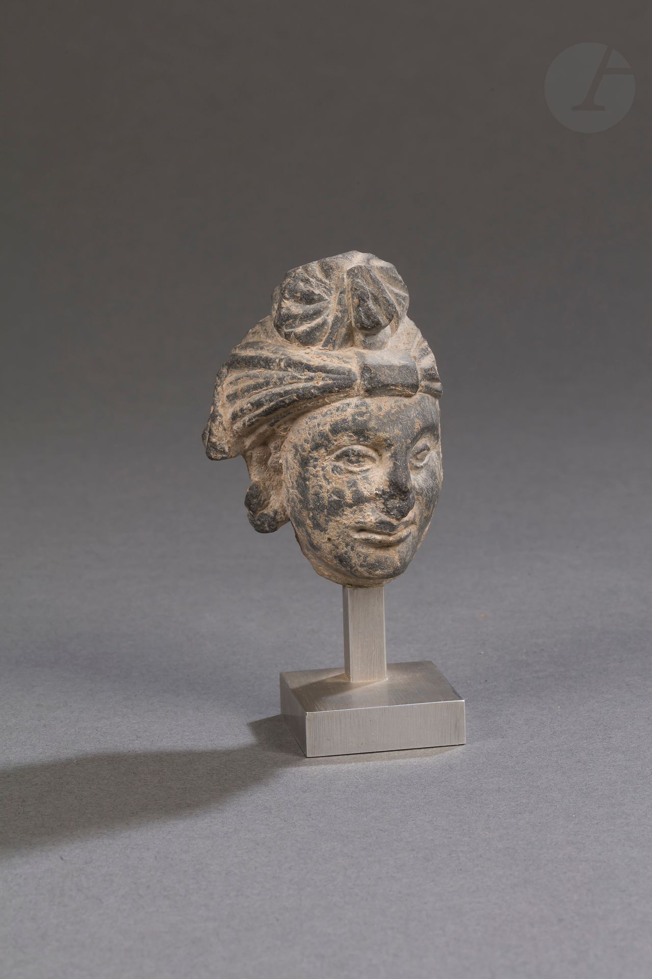 Null 深灰色片岩小佛头，希腊佛教艺术，犍陀罗，2-5世纪
头发被缠住，在头骨的顶部聚成一个发髻。金属底座。
高度：7.5厘米