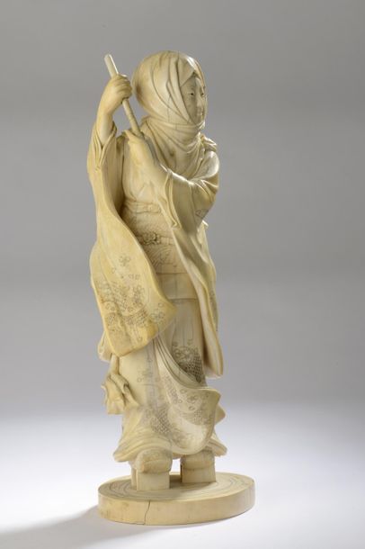 null Okimono en ivoire sculpté et rehaussé à l’encre, femme debout sur de hautes...