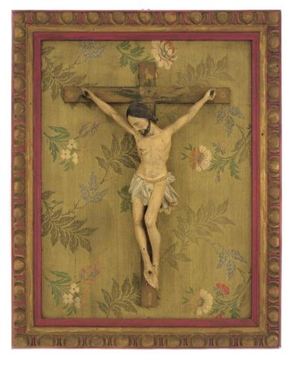 Christ en croix en bois polychrome reposant dans un cadre sur fond d'un tissu fleuri
