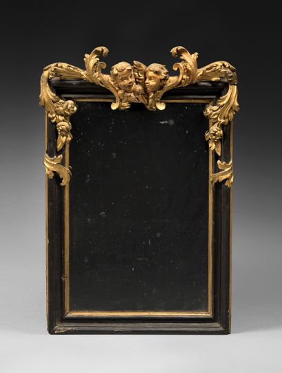 null A BAROQUE MIRROR with a blackened and gilded wood frame, topped by two cherubs'...