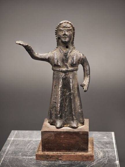 青铜小雕像，刻画了一位风格化的站立女性形象，右臂高举。

面部以简笔画手法表现，发型环绕脸庞。身体穿