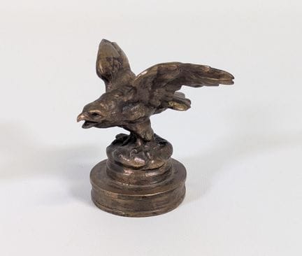 一件青铜錾刻汽车装饰品，表面呈金棕色古铜色，造型为展翅的雄鹰，底座上署有“Antonio DUART