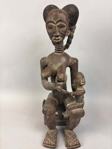一件表面呈深色古铜色的木雕母性雕像，刻画了一位坐姿哺乳的妇女，颈部修长且带有环状纹路，头顶盘起高高的
