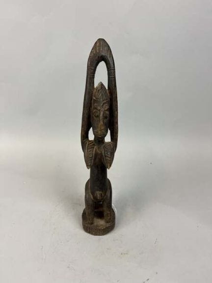 一件表面呈深色包浆的木雕人形雕像，刻画了一位双臂高举过头的人物，头戴带有锯齿状饰纹并刻有纹路的头饰，