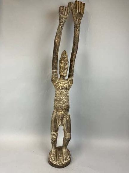 一尊木雕雕像，双臂高举，表面呈褐色包浆，刻画了一位双臂高举的站立男性形象，躯干和四肢饰有刻划的几何图