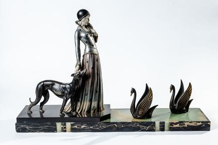 这组雕像采用古铜色和银色釉面处理，描绘了一位优雅的女士携猎犬，身前有一对天鹅。底座由大理石和缟玛瑙制