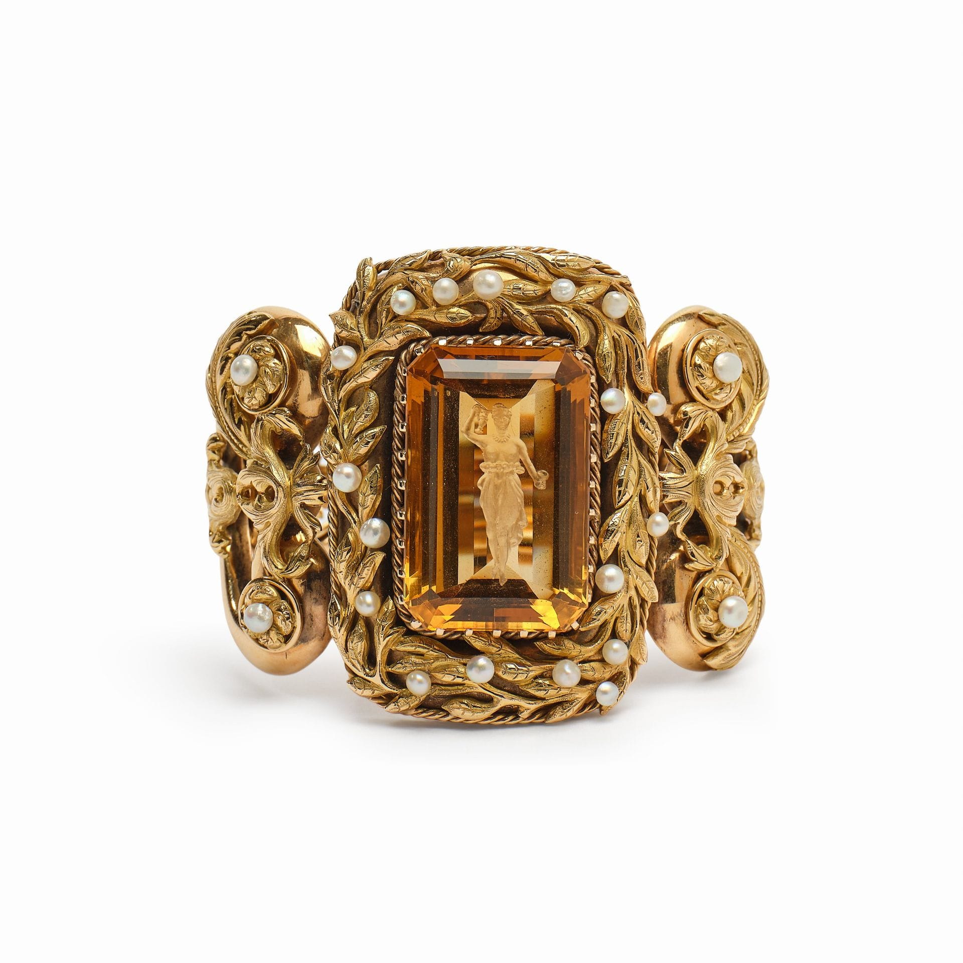 IMPORTANT BRACELET EN OR ET CITRINE, TRAVAIL ITALIEN DU XIXE SIÈCLE
en or jaune le motif central serti d'une citrine rectangulaire à pans coupés gravée en intaille d'une figure d'Hébé, dans un entourage de feuillages ciselés ajouré et appliqué semés de perles ; les éléments latéraux sont ajourés de volutes affrontées à décor de palmettes, feuilles d'acanthes et rosaces centrées de perles. 
Travail italien du milieu du XIXe siècle 
Non signé
Haut. 6 cm - Larg. 7 cm - Prof. 7 cm
Poids brut : 124,5 g

La table de la citrine est gravée en intaille d'Hébé. La déesse est représentée debout, le buste nu, les cheveux relevés et parée d'un collier. Une ceinture fleurie retient son drapé, qui s'envole en plis souples. D'une main elle élève une aiguière, de l'autre elle tient une coupe.
Fille de Zeus et d'Héra, Hébé personnifie la jeunesse éternelle. Échanson de l'Olympe, elle versait aux dieux le nectar et l'ambroisie, d'où ses attributs, l'aiguière et la coupe. Après l'apothéose d'Héraclès, elle devint son épouse.
Le thème connut une grande faveur au XIXe siècle, en particulier en Italie, après les célèbres marbres d'Antonio Canova (1757-1822). La figure du bracelet s'inscrit dans cette tradition néoclassique.