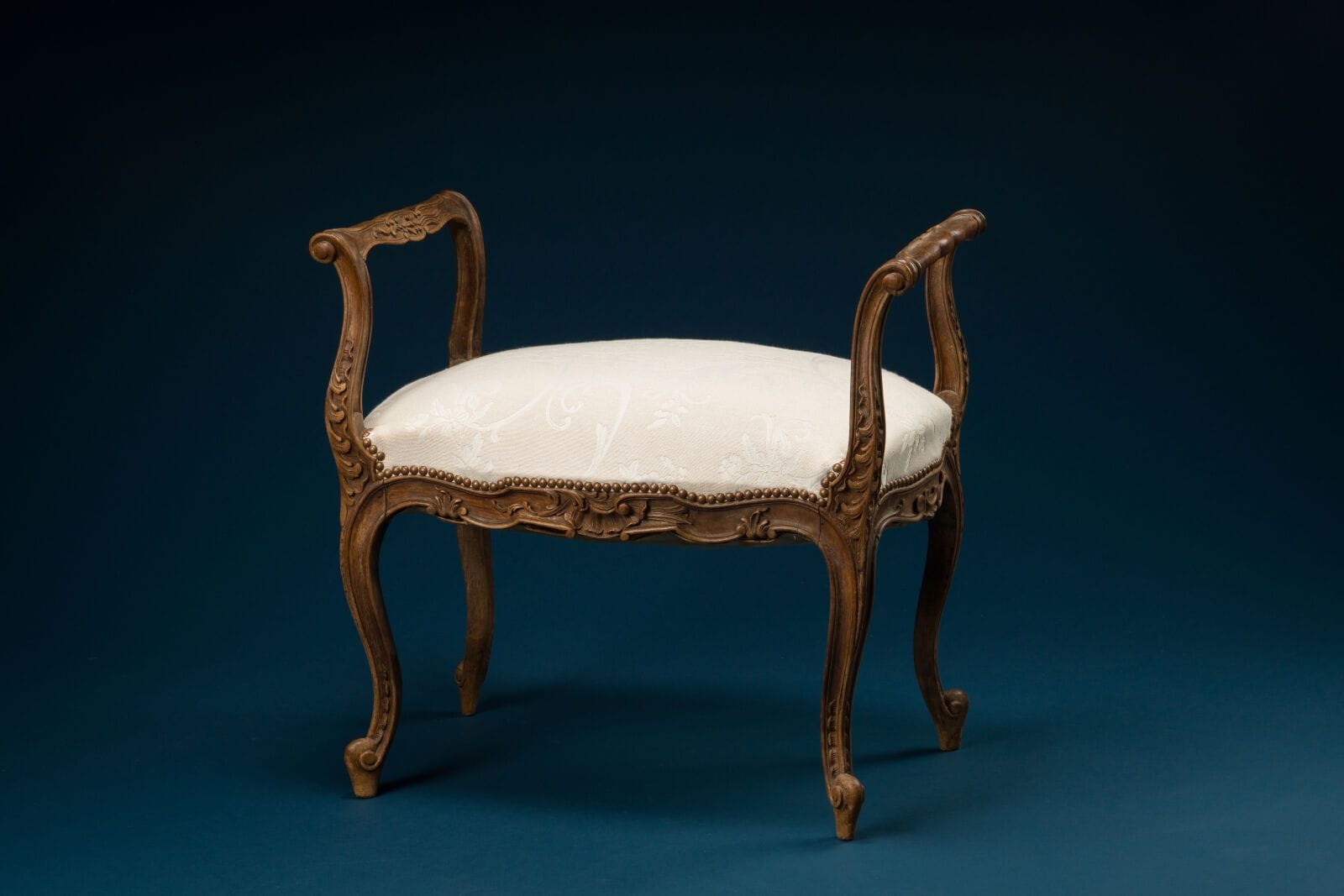 Small bench with armrests carved from natural wood in a floral motif, curved legs, and white fabric upholstery
Louis XV style.
H.: 66 cm; W.: 69 cm; D.: 38.5 cm.