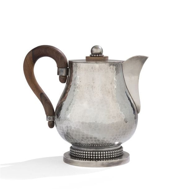 JEAN DESPRÉS (1889-1980) 
Élégante verseuse à café ou à thé à une anse détachée en palissandre. Épreuve en métal argenté. Le corps du verseur, achevé en martelage, repose sur un pied orné d'un triple rang perlé et reçu par une base circulaire traitée lisse. Le couvercle, également traité lisse, reçoit la graine sphérique reposant sur un rang perlé puis sur une platine circulaire en palissandre. Signé J. DESPRÉS à la pointe sous la base et porte, à deux reprises, le poinçon de Maître Orfèvre sur la tranche de la base. H. 18,5 cm