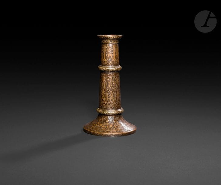 Petit flambeau, mash’al, en laiton, Iran qâjâr, XIXe siècle 
Fût cylindrique scandé de deux bandeaux saillants sur base évasée, à décor de médaillons de divs et de créatures hybrides sur l’ensemble. à la base, frises d’arabesques de palmettes bifides. 
Hauteur : 19,5 cm 

A Qajar brass candlestick, mash’al, Iran, 19th century