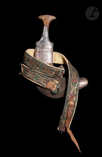 Poignard djambiya, région d’Oman, début XXe siècle 
Lame courbe à double tranchant renforcée d’une nervure médiane. Monture en corne rehaussée de garnitures en argent. Fourreau en bois recouvert de velours brun sur une face, cousu de fils d’argent sur l’autre et garni de plaques d’argent ciselé aux extrémités. Anneaux d’attache latéraux à une ceinture en tissu broché de fils colorés et dorés dessinant une frise végétale ponctuée de losanges. Une pièce iranienne en argent de 50 rials fixée sur un pan. 
Longueur avec fourreau : 29,5 cm ; 
Poids brut : 520 g
État : chocs sur la monture, rayures sur la lame, ceinture usée. 

Bibliographie :
Alain Jacob, Les armes blanches du monde islamique, Paris : 1985, pp. 138-139. 

A dagger, djambiya, Oman region, early 20th century