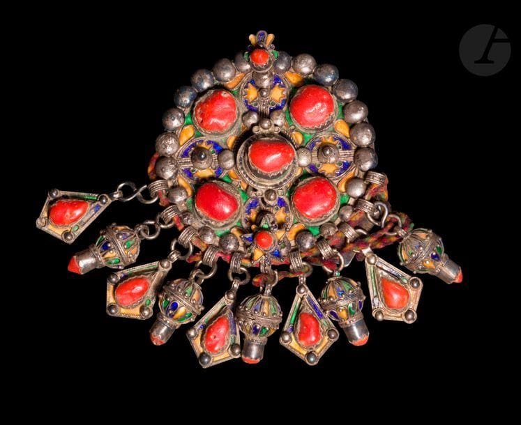 Grande fibule ronde, tabzmit, Algérie, Grande Kabylie, Beni Yenni, début XXe siècle 
Fibule ronde, en argent couvert d’émail cloisonné tricolore bleu, vert et jaune, sertie de cabochons de corail et bordée de perles appliquées. Au revers, étoile à huit branches émaillées et en partie inférieure, 11 pendeloques de deux types, émaillées en polychromie et à cabochons de corail. 
Diamètre : 10,5 cm ; poids brut 337,5 g 
État : ardillon manquant, irisation, encrassement, liens de laine et de textile ajoutés.

An enameled silver fibulae, Algeria, Great Kabylia, Bani Yenni, early 20th century 