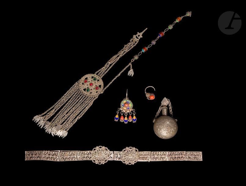 Ensemble de bijoux en métal argenté, Afrique du Nord, XXe siècle 
Une ceinture en plaquettes à décor floral ajouré, longueur : 72,4 cm ; un flacon à khôl globulaire à décor végétal ciselé et chaîne de suspension, hauteur avec bouchon : 10 cm ; un petit bracelet à perles en argent émaillé et en cornaline, longueur avec fermoir : 19 cm ; une boucle d’oreille à décor émaillé et cabochon de corail, longueur : 2,9 cm ; une petite fibule à décor émaillé et cabochons de corail, hauteur : 7,5 cm ; un ornement de coiffe circulaire serti de pâtes de verre colorées et encadré de chaînes, quatre de suspension, seize en pendeloques achevées par des mains stylisées. Longueur : 33 cm
État : ardillon de la fibule tordu, traces d’oxydation sur l’ornement de coiffe.

A lot of jewels, North Africa, 20th century 