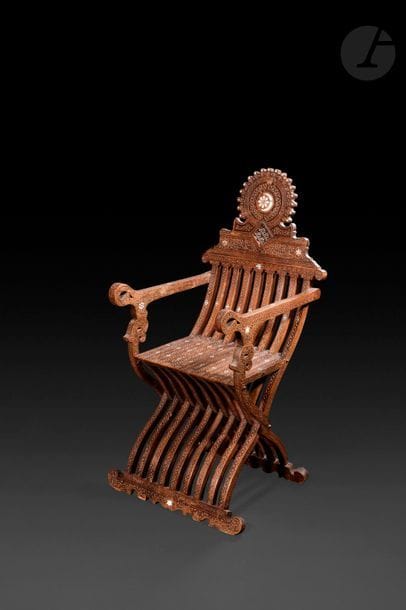 Fauteuil pliant en bois foncé, Empire ottoman, Syrie, XIXe siècle 
En bois sculpté et incrusté de nacre et d’ébène, à dossier surmonté d’une rosace, accoudoirs droits. Sur l’ensemble, décor sculpté de rinceaux et de palmettes rehaussé de motifs étoilés et inscriptions en thuluth sur le dossier.
114 x 56 cm ; siège : 45 x 35,5 cm 

A folding wooden chair, Ottoman Empire, Syria, 19th century