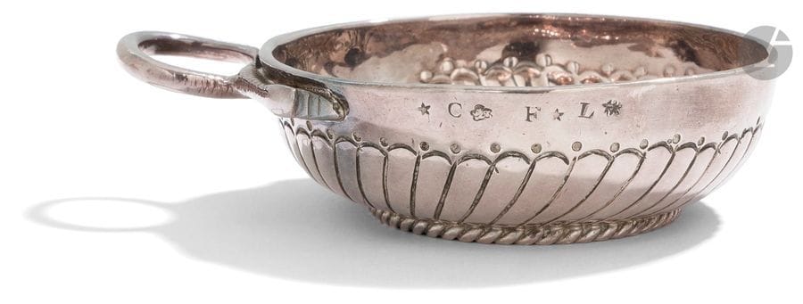 PARIS 1750 - 1756
Tasse à vin en argent, elle repose sur une bâte cordée, le corps est ciselé de côtes torses et rangs de points, anse serpent non poinçonnée. Elle porte en son centre une pièce à l’effigie du Roi Louis XV datée de 1756. Gravée des lettres F et L sur le pourtour. 
Poinçon de décharge au sanglier. 
Poids : 137,20 g - diamètre sans l’anse : 8,9 cm