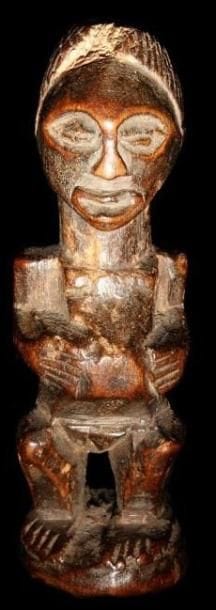 Statuette fétiche SONGYE (R.D.Congo). Personnage féminin debout sur un petit socle cylindrique, les mains ramenées vers un ventre proéminent recevant une charge magique. Cou puissant supportant une tête au visage souriant, charge sur le sommet du crâne. Sculpture très équilibrée à la patine brun nuancé brillante et de grande ancienneté.. Hauteur:9 cm.