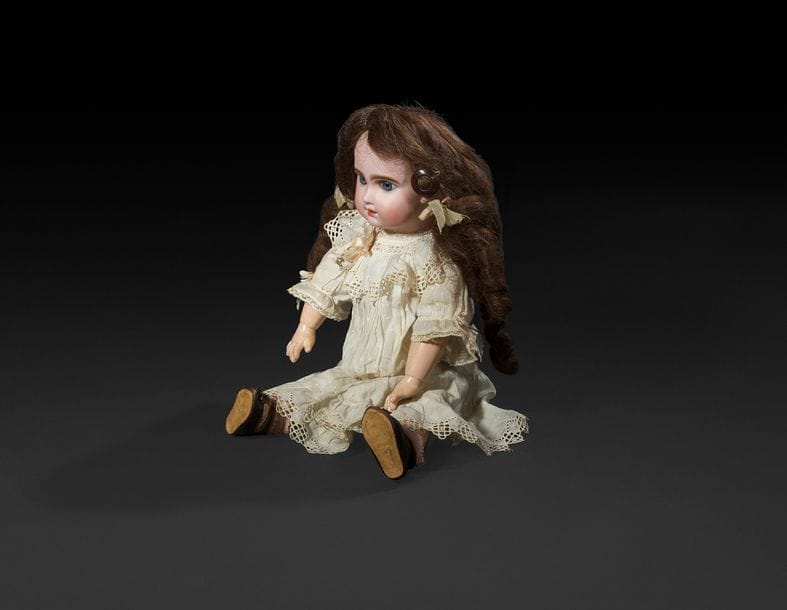 Bébé JUMEAU, tête en porcelaine marquée au tampon rouge : Tête JUMEAU, T. 7, yeux fixes bleus, bouche ouverte sur une rangée de dents, bouche ouverte sur une rangée de dents, oreilles percées, perruque brune. Corps articulé en bois et composition, étiquette bleue : Bébé JUMEAU Diplôme d’honneur, robe blanche à pois, souliers d’origine marqués à l’abeille, T.7.
H : 45 cm