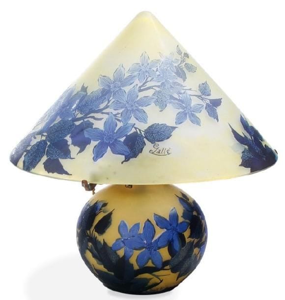 Émile GALLE (1846-1904)
Abat-jour de lampe en forme de chapeau chinois Épreuve de tirage industriel en verre multicouche dégagé à l'acide à décor, bleu parme, de branches fleuries de pommier sur fond jaune opalescent. Signature en réserve gravée en camée à l'acide. Hauteur: 20 cm - Diamètre: 36,5 cm. On y joint un pied assorti pour recomposer une lampe.