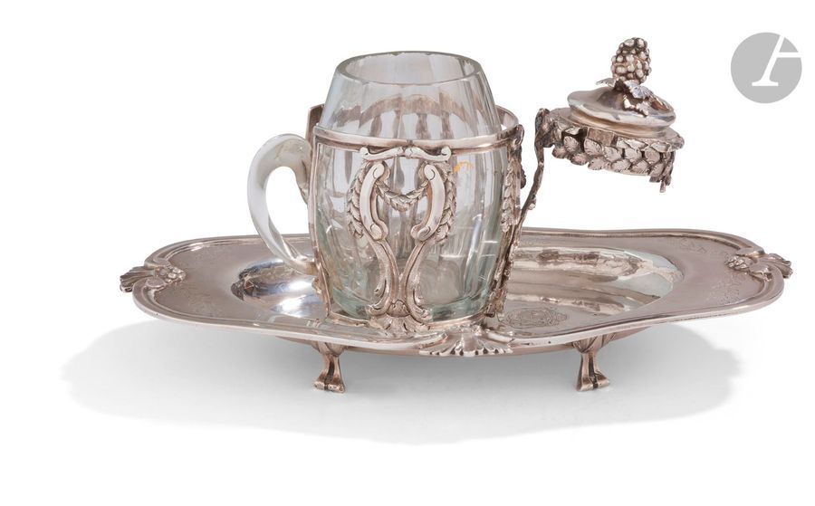 TOULOUSE 1777
Moutardier en argent et verre (traces de dorure) posé sur quatre pieds à attaches feuillagées, et un couvercle portant la même décharge (un oiseau), vissé sur un plateau de forme ovale chantournée, mouluré de filets, rehaussé d’agrafes de feuillages et de fruits et ciselé de guirlandes feuillagées. Gravé d’armoiries d’alliance surmontées d’une couronne de marquis et d’une toque de président à mortier. 
Maître orfèvre : Louis II DULAURIER, reçu en 1765
État : manques des feuilles de vigne sur la prise du couvercle.
Poids net : 324,20 g - Longueur du plateau : 20,5 cm