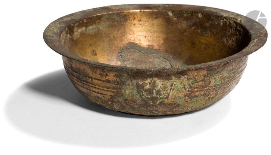 CHINE - Époque HAN (206 av. JC - 220 ap. JC)
Bol en bronze à traces de dorure à bordure évasée, la panse ornée d’un ourlet, les côtés flanqués de deux anses formant têtes de taotie. (Petits manques et restauration).
Diam. 16,9 cm