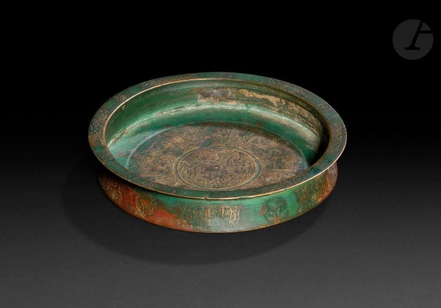 Bassin en bronze, Iran oriental, Khorassan, XIIIe siècle, montage XIXe siècle
Bassin cylindrique peu profond à fond plat et large lèvre plate et à décor gravé. Sur la panse, alternance de cartouches épigraphiques en calligraphie coufique et de médaillons circulaires à décors végétaux, sur la lèvre, frise tressée interrompue par des médaillons circulaires, et dans le fond, figure centrale de sphinge entourée de caractères cursifs sur fond de rinceaux végétaux.
Diamètre ouverture : 17,8 cm ; hauteur : 3,5 cm
État : restaurations.

A shallow bronze bowl, Eastern Iran, Khorasan, 13th century, remounted 19th century