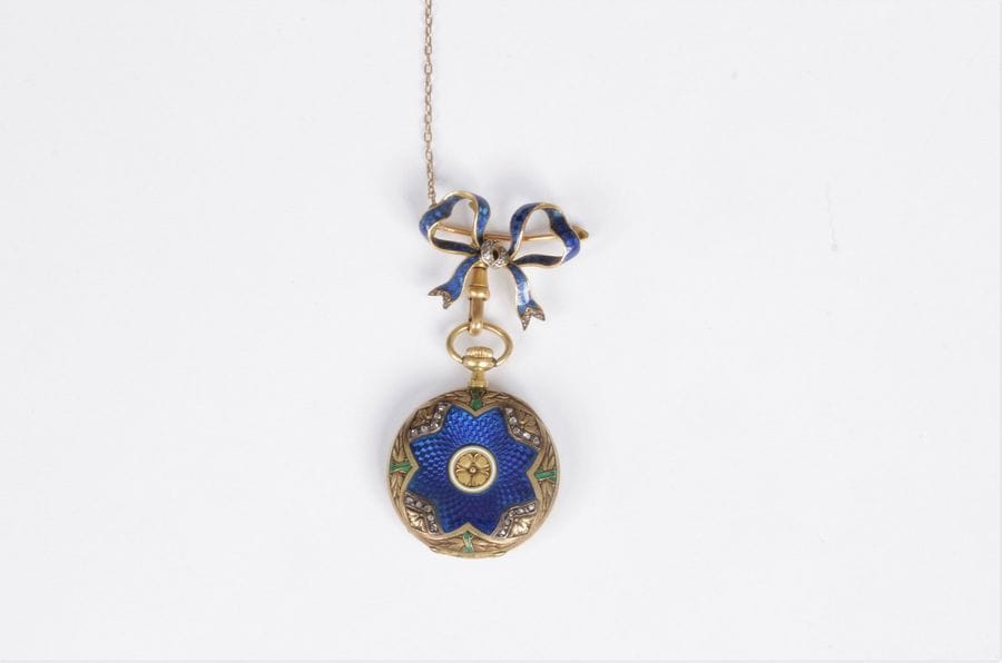 Montre de col en or 18K (750), le fond émaillé bleu sur motifs guillochés centrés d’une fleur ciselée, les bords agrémentés de diamants taillés en roses, retenue par une broche dessinant un nœud. Dans un écrin de la Maison Mellerio. Travail français de la fin du XIXe siècle. Diamètre : 2,6 cm environ. Poids brut : 24,6 g