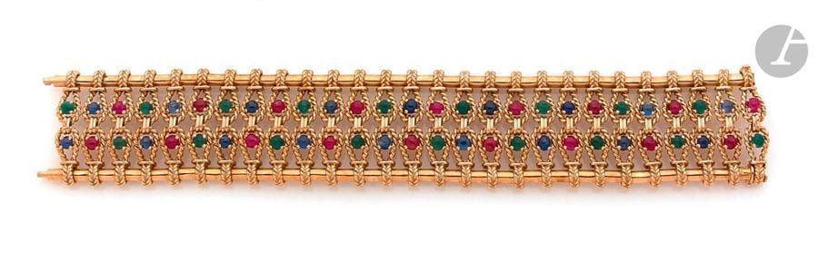 Bracelet large articulé en or 18K (750) cordé et lisse, ponctué de rubis, de saphirs et d’émeraudes cabochons. Travail français des années 1960. Dimensions : 19,5 x 3,2 cm environ. Poids brut : 101,1 (égrisures)