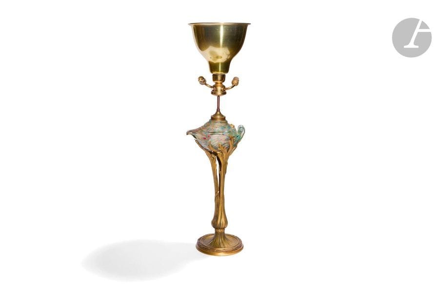 JOSEPH-FRANÇOIS JOINDY (1832-1906) SCULPTEUR & GEORGES GAGNEAU FONDEUR-ÉDITEUR
Roseaux et lampe d’Aladin, le modèle présenté au Salon de la Société des Artistes Français, Paris, 1898
Importante lampe à huile naturaliste électrifiée postérieurement. Le pied (base et fût) en bronze doré. Le réservoir en verre aux colorations polychromes intercalaires évoquant une lampe d’Aladin et dans le goût des verreries d’Ernest-Baptiste Léveillé. Circa 1900. Un petit éclat dans le verre en partie basse et masqué par la monture, électrification non d’origine, un enfoncement en pourtour de la base. H. totale avec système électrique : 88 cm - H. sans système électrique : 52 cm

Bibliographie :
Alastair Duncan - The Paris Salons 1895-1914- Volume V : Objets d’art & Metalware – Édition Antique Collectors’ Club, 1998. Modèle identique reproduit page 331.