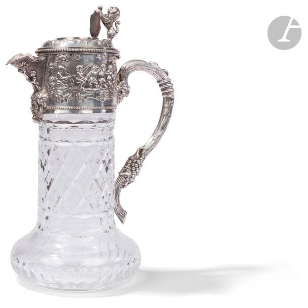 PORTUGAL - PORTO XXe siècle
Une aiguière en cristal taillé et monture en argent 925 ‰. De forme circulaire, le cristal reçoit dans sa partie supérieure une frise repoussée sur deux faces de scènes figurant des putti dansant et jouant encadrant un bec verseur souligné d’une tête de Bacchus, et une anse à l’imitation d’un cep de vigne. L’appui-pouce du couvercle à charnière figure un lion héraldique tenant un blason dont le champ est laissé vierge de gravure.
Orfèvre : TOPAZIO
État : la monture à refixer
Poids brut : 1302,84 g - hauteur : 28,5 cm - diamètre de la base : 13,5 cm