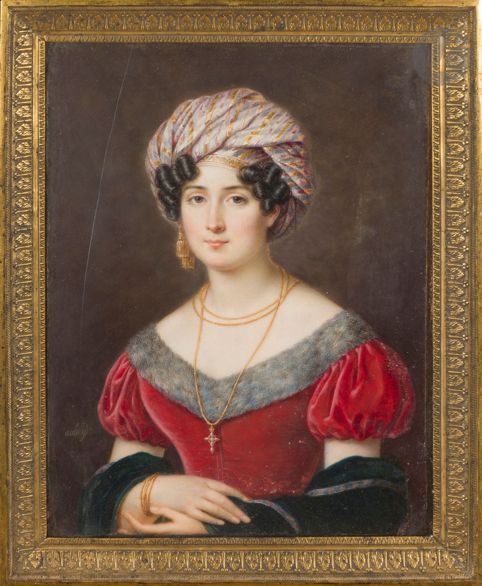 Louis François AUBRY (1767-1851) 
Portrait présumé de Julie Candeille (1767-1834), actrice, musicienne, femme de lettres et femme d’affaires, âme sœur du peintre Girodet. Elle est vue de trois-quarts à droite, en robe rouge à manches courtes bouffantes et col d’hermine, et grand sautoir avec croix en or. Elle est coiffée d’un turban à rayures dorées découvrant des cheveux bruns bouclés sur les tempes. Ses mains croisées devant sa taille laissent entrevoir l’étole de velours bleu couvrant ses avants bras. 
Grande miniature sur ivoire 
18,5 x 14,5 cm
Signé en bas à gauche Aubry 
Fente et restauration 
Cadre en velours marron, frise de palmettes en laiton doré 

Julie Candeille, « ce joli monstre de talents » selon les frères Goncourt, posa avec le même turban mais en robe blanche à col de dentelle, pour une gravure vers 1810 de Sébastien Coeuré d’après Prud’hon. 
Louis François Aubry, l’un des meilleurs miniaturistes de son temps, forma de nombreux élèves. Le musée Antoine Lecuyer de Saint Quentin (Aisne) conserve une copie d’atelier de notre miniature, vue sans les mains et le châle, le modèle non identifié dans les inventaires (inv. 2006.28.1FA).

Un même portrait, non identifié, signé Mme de Mirbel a figuré dans la vente Pierre Bergé et Associés, 21 décembre 2006, 19,6 x 14 cm avec cadre.
