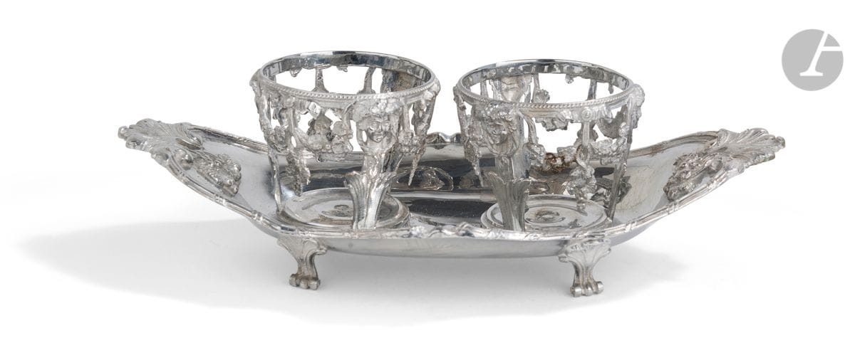 Huilier vinaigrier en argent fondu de forme ovale sur un modèle de la fin du XVIIIe siècle. Elle repose sur des pieds à enroulement à attache de coquille, le pourtour est rehaussé de filets et rubans croisés associés aux extrémités de feuillages et de grappes de raisin. Les porte flacons alternent personnages en terme et grappes de raisin en chute surmontés de nœuds. 
Poinçon minerve
Poids : 790 g - longueur : 30,2 cm - largeur : 15,7 cm