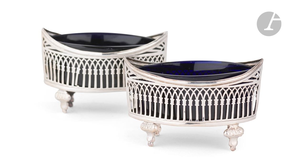 PARIS 1798 - 1809
Paire de salières en argent estampé et deux verres bleus de forme ovale quadripode, à décor ajouré d’arcatures. 
Poinçon au premier coq
Orfèvre : Ambroise MIGNEROT, insculpe en 1 800, biffé en 1818
Poids net : 59,9 g - longueur : 9,5 cm - largeur : 4,3 cm
État : une arcature à ressouder